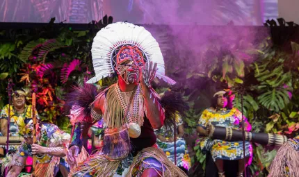 Dancer dressed in traditional Torres Strait Islander ceremonial clothing in the foreground with musicians in similar wear in background.