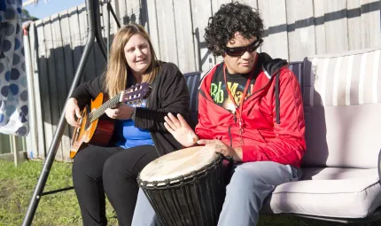 Seated on a large swing chair, an Indigenous woman dressed in black plays a guitar while an Indigenous man in red top and grey pants plays a large bongo drum.