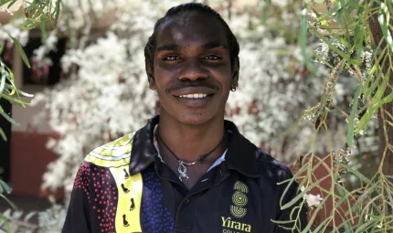 A young man in a polo shirt with traditional designs smiles at the camera. A tree branch with leaves and flowers hangs in front and to his left. In the background is more foliage.