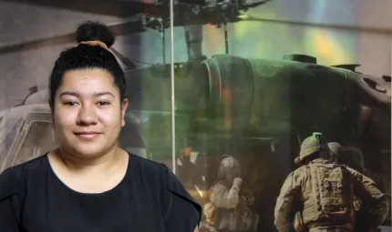 A woman wearing a black shirt with her hair in a knot. In the background is the image of a helicopter with military personnel in front of it.