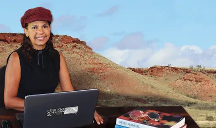 A woman in black top and red cap sits at a desk on which is a laptop and two books. In the background is a large photo of a dry landscape comprising two hills ochre red and pale green in colour.