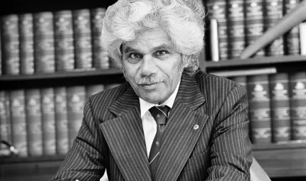 Elderly man with grey hair wearing a striped suit sits in front of a shelf lined with many bound books.