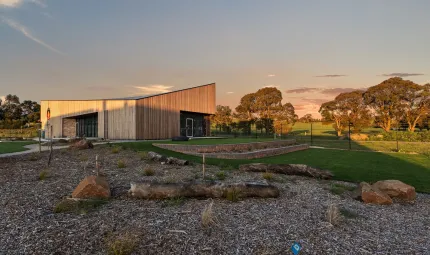 A building with two entries at left and right with wooden walls and a sloping roof is surrounded by grass and wood chip. In the background are trees and pastures.