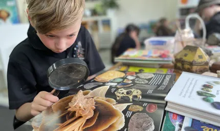 A boy with light-brown hair is looking through a magnifying glass at an object. Underneath the object is a large textbook about rocks. The bench is scattered with books and other objects.