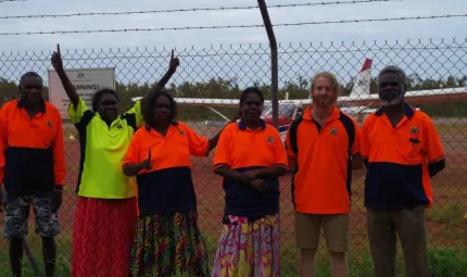 Five Aboriginal people and one non-Aboriginal person are standing in front of a remote airfield. In the background is a small airplane.