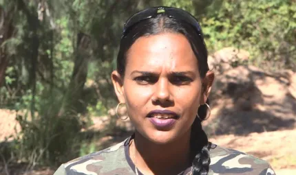 Aboriginal woman in cargo shirt and hair in plat looks at camera with trees in the background.