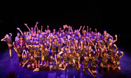 A large group of youth and a few adults stand or sit on a purple-lit stage. The background is black.