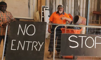 Two men in workwear stand behind signs leaning up against a gate. The signs say ‘No entry’ and ‘Stop’. In the background is a shed wall.