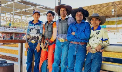 Five boys stand side by side leaning against a wooden fence. Behind them is a rodeo ring. Each of the boys is wearing jeans and a belt or work pants and collared work shirts. The boy on the left is wearing a cap and four of the boys aren't.