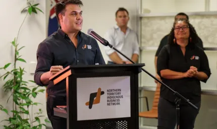 One person, dressed in black shirt with NTIBN logo is the focus of this image.  They are speaking into a microphone behind a lectern which displays a sign showing a logo which reads “Northern Territory Indigenous Business Network”.