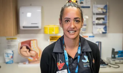 A woman in a nurse’s uniform with hair tied back faces camera. In the background is a bench with apparatus on it and a wall with more items attached to it.