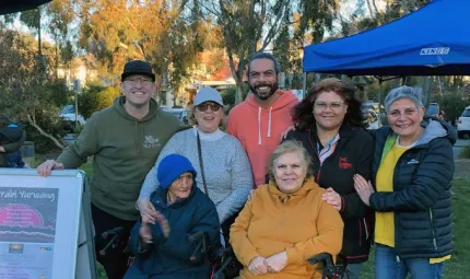 7 adults stand in a close group outdoors in front of a blue marquee tent. Each of them has their hand on the shoulder or arm of each other. The man on the left has his hand on a whiteboard sign that has a poster for Yerrabi Yurwang.