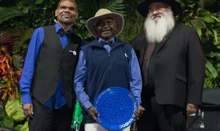 An elderly Aboriginal man holds a large blue plate with a younger Aboriginal man standing to his right and an Aboriginal man with a grey beard standing to his left.