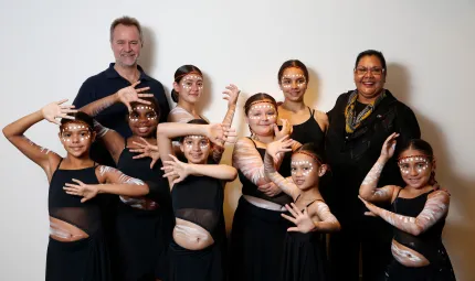 Non-Indigenous man and Aboriginal woman stand in front of white background with young female Indigenous dancers between and in front of them. All are dressed in black and the dancers wear white face paint.