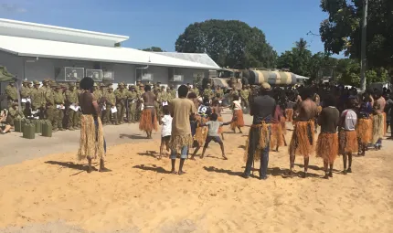 A group of dancers in grass skirts dancing on sand for a group of army soldiers wearing green fatigues.