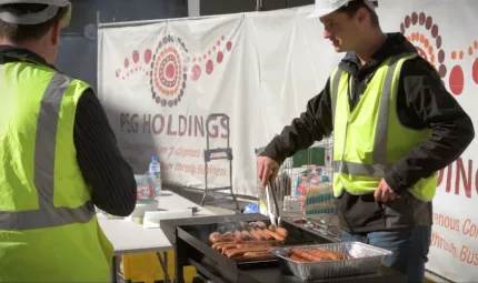 Man in yellow high visibility vest and white hard hat turning sausages on BBQ in front of PSG Holdings site and a building site. 2nd man on right side in high vis vest with back turned.