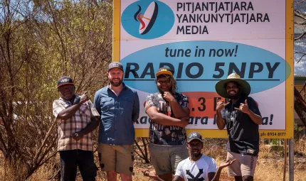 Five men in casual wear stand or sit in front of a large multi-coloured sign with a logo at top left and the following text: Pitjantjatjara Yankunytjatjara Media. Tune in now Radio 5NPY. In the background are trees, grass and a building.