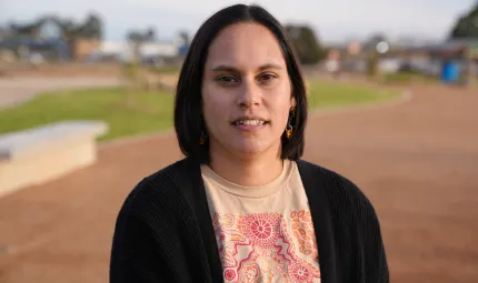 A woman with dark shoulder length hair and wearing a black top over a colourful top with traditional designs faces camera. In the background is brown soil, green grass and some buildings.