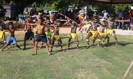 A group of Indigenous children with white ceremonial paint on their bodies wearing shorts or yellow coverings dance on green grass watched by a crowd of onlookers.