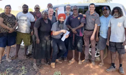 There are 12 adults standing together and facing the camera. They are outside on a sunny day, standing in the shade, in front of a white van.