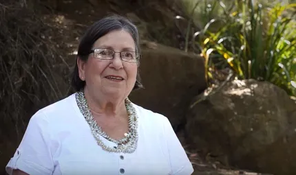 Elderly woman with dark hair wearing glasses, multiple necklaces and white dress stands in front of rocks and green foliage.