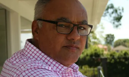 A man wearing a red checked shirt and black rimmed glasses looks at camera. In the background are bushes, a fence and some grass. Above him is a ceiling.