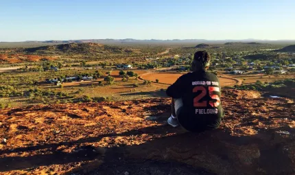 Aboriginal woman sits on a hill overlooking a small town surrounded by low hills in an arid landscape. On the back of her dark top is ‘Squad of 2017, 25, Fielding’.