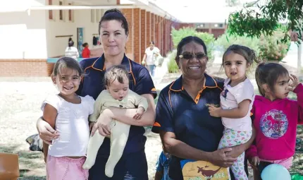 Two Indigenous women in dark blue shirts hold or support four Indigenous children, the group stand in front of a brick building mostly painted white with trees nearby.