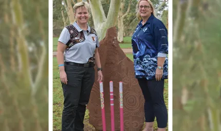 Two women stand either side of a rock slab sculpture with Indigenous designs in front of which are three cricket stumps coloured pink and decorated with colourful Indigenous designs. The woman on the left wears dark trousers and a grey and brown shirt.