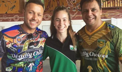 Two men and a young woman who stands between them face the camera. They all wear colourful shirts and in the background is a wall painted with Indigenous designs.