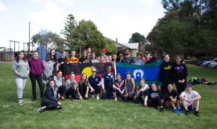 Group of Indigenous youth sit or stand on grass, some holding the Aboriginal flag and Torres Strait Islander flag. In the background is a building, cars and trees.