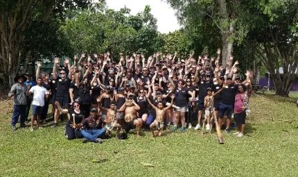 A huddled group of staff in Yarrabah, covered in Indigenous paint, standing with their hands in the air. Some staff are holding Indigenous instruments.