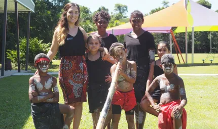 An adult woman and seven Indigenous youth stand or kneel on a lawn. To the left is a building and in the background a canopy. Some of the youth wear paint in traditional patterns and wear traditional clothing while one holds a didgeridoo.