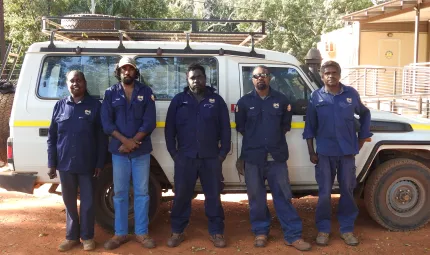 An Aboriginal woman and 4 Aboriginal men all dressed in blue ranger uniforms stand on red soil and in front of a white 4 wheel drive vehicle. In the background is a building and trees.