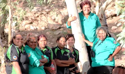 Seven Aboriginal women in ranger uniforms stand next to a white bark tree. In the background is a rocky slope and more trees.