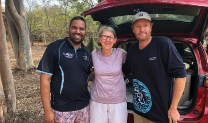 A mature woman in lilac coloured top and white shorts stands between, and arm in arm, with two men dressed in blue shirts. Behind them is a red car with its rear door open. In the background are trees and bare soil.