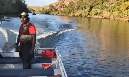 A young man stands up and steers a boat on a river. In the background is water, vegetation and a cliff face.