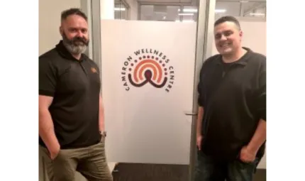 Two men in dark shirts and trousers stand either side of a door which has a sign on it. The sign features the words Cameron Wellness Centre partially wrapped around an Aboriginal dot and line design.