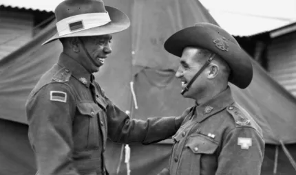 A smiling man in army uniform shakes hands with another smiling man also in uniform. In the background is a large tent and a building.