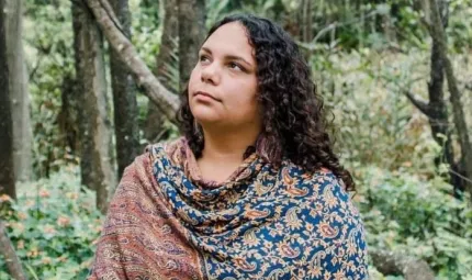 A woman with long dark hair wearing a colourful and patterned top stands in a forest. In the background are trees and shrubs.