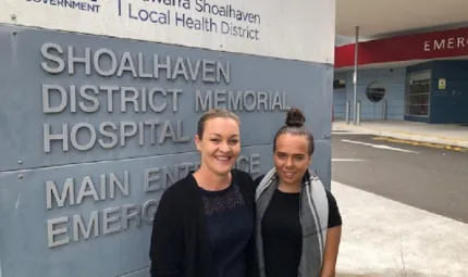 Two women stand in front of a large sign featuring the words Shoalhaven District Memorial Hospital. The woman at left wears a dark cardigan and dress and the one on the right wears a dark dress with a grey and white scarf about her neck.