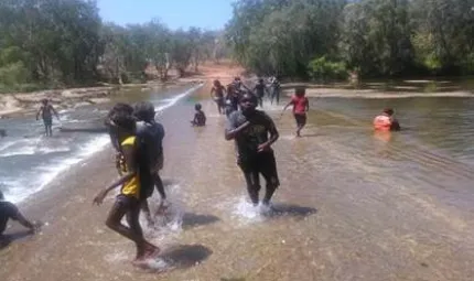 Several youth stand on a roadway over which flows a river. At left are rocks in the stream and in the background is a dirt road, trees and blue sky.