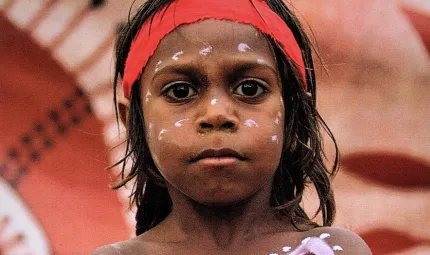 Aboriginal boy in body and face paint standing in front of Aboriginal artwork