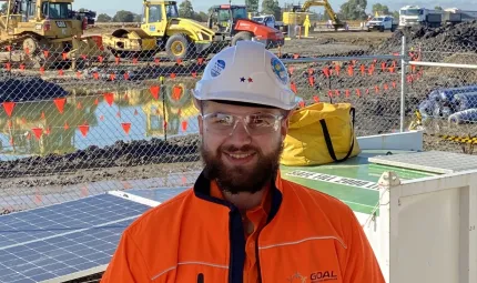 A young adult male in orange work-wear and white helmet stands in front of a white container and solar panel. In the background are large earthmoving vehicles and trucks.