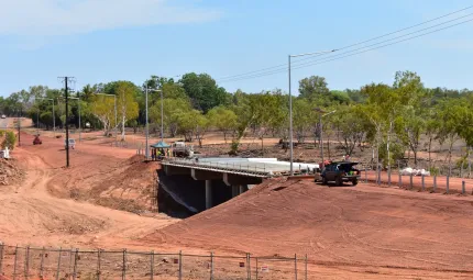 In the foreground is ochre coloured soil behind which is a concrete bridge with vehicles nearby and in the background are trees and blue sky.