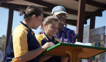 Two young Aboriginal girls dressed in blue and yellow polo shirts stand at a podium with an elderly Aboriginal man in blue cap and blue and red shirt. In the background is a small metal shelter.