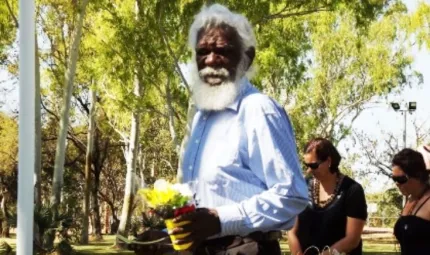 Elderly Aboriginal man with white hair and beard and dressed in pale blue shirt is holding a pot of flowers. In the background are two women dressed in black and a line of tall leafy trees.