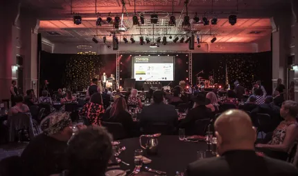 Large group of Aboriginal and Torres Strait Islander people sitting at tables in a darkened room. In the background is a stage and on the ceiling, stage lights.