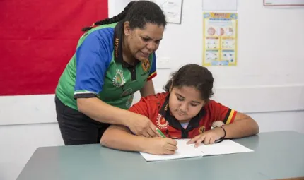 An Indigenous teacher in a green polo shirt helps an Indigenous student in a red polo shirt with their school work.