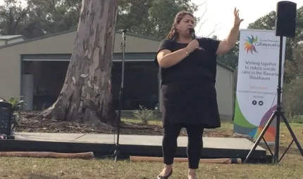 Aboriginal woman in black clothing holds a microphone as she stands on grass. In the background is a large tree, a shed, a sign, a speaker on a stand and some didgeridoos laying on the grass.